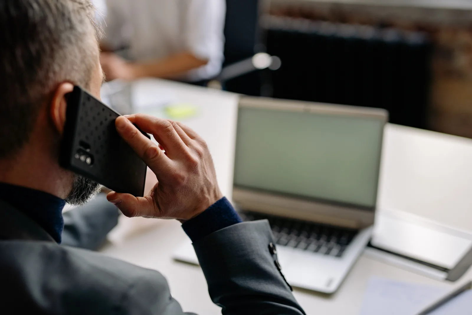 Professional man using cellphone in office setting, multitasking with a laptop. Business email compromise: stop payment fraud by phone