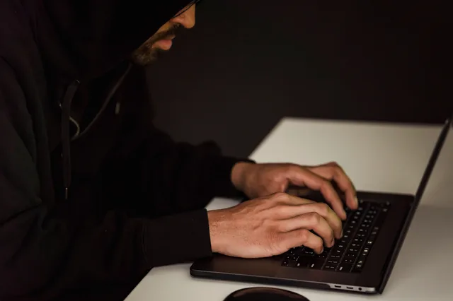 Crop bearded male hacker in black hood typing on netbook keyboard and browsing dark net in obscure room. Business email compromise comparison: what to keep and what to drop